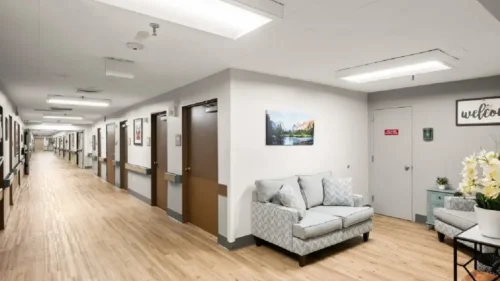 A hallway with wood floors and a seating area with a gray couch, a light blue armchair, and a landscape painting
