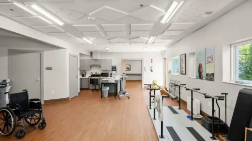 A physical therapy room with a parallel bar track, exercise equipment, and a kitchenette area in the background