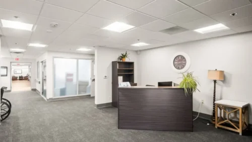 A reception area with a dark wood desk, a large wall clock, and a gray carpeted floor, leading into a hallway