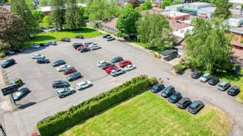 An aerial view of a large parking lot filled with cars, a grassy area, and several buildings surrounded by tree